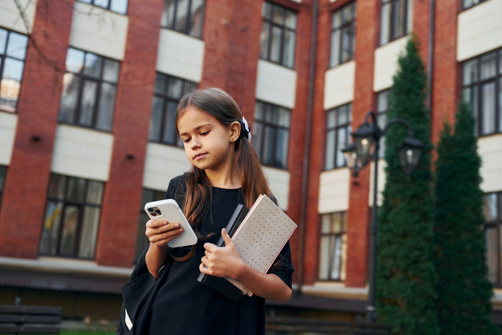 Holding phone in hands. Schoolgirl is outside near school building